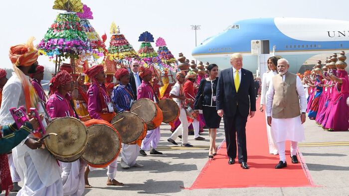 To menn på en løper på foran et fly i India - to kvinner bak  Det er trump og Modi i front. Og masse indere med trommer og fargerike klær som tar imot dem. 