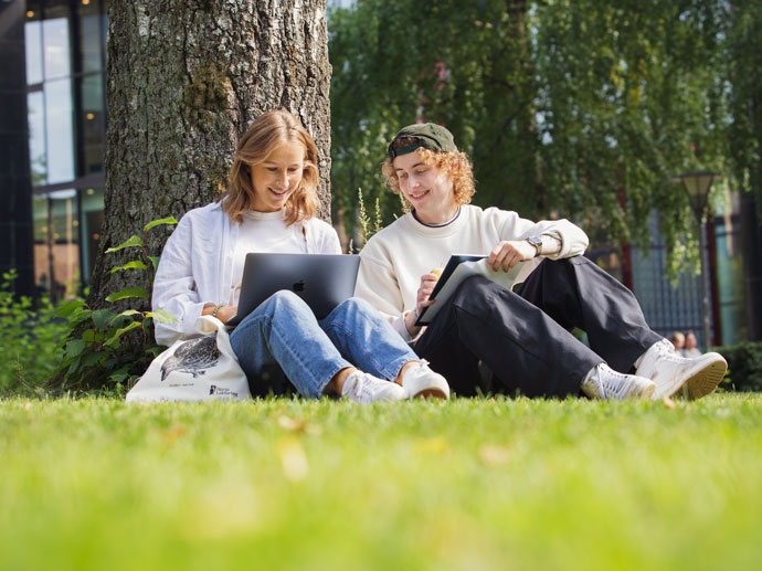 Two students sitting on a lawn by a tree with their laptops