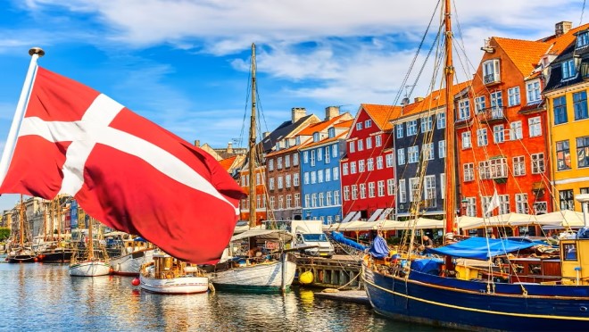 Danish flag in the foreground with a dock with boats and colorful buildings in the background
