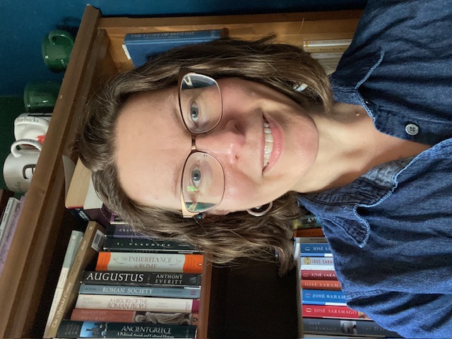 A smiling, caucasion woman with shoulder length, wavy hair, wearing glasses. She is sitting in front of a book shelf, loogking into the camera. 