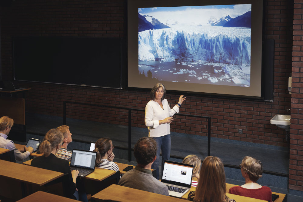A teacher and her students sitting in an auditorium, presenting a picture of a glacier in the background.