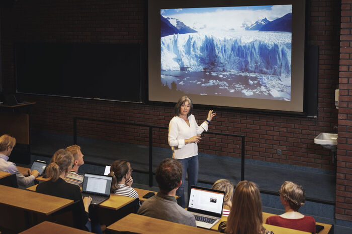 A teacher and her students sitting in an auditorium, presenting a picture of a glacier in the background.