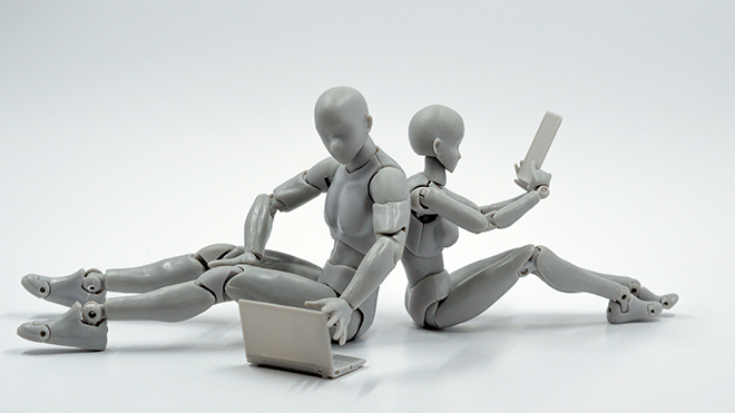 To mennekselignende figurer laget av lys grå plast sitter rygg mot rygg, en med plast-laptop og en med plast-nettbrett. Fotografi.