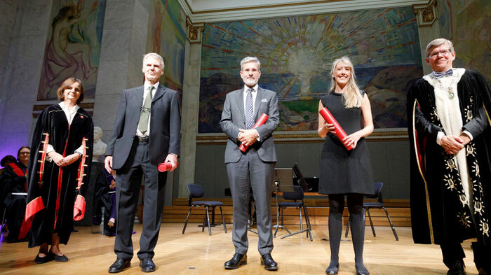 Fem voksne personer ser mot kamera med en rød rull i hendene inne på scenen i aula med Much-malerier bak.
