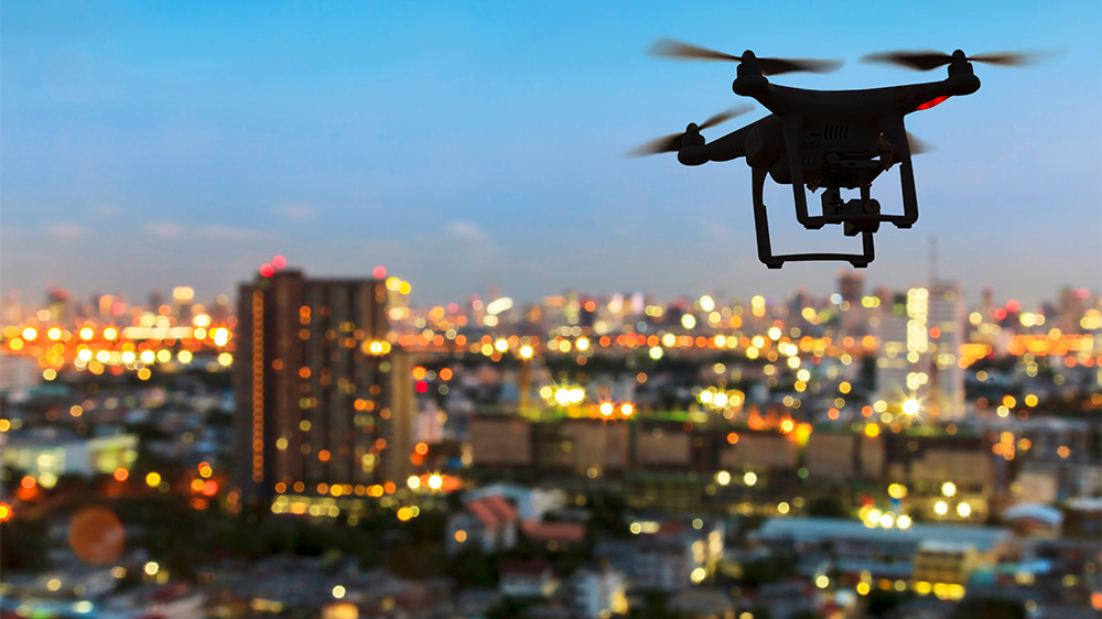 Silhuett av drone til høyre i luftrom på kvelden over en lyssatt by.