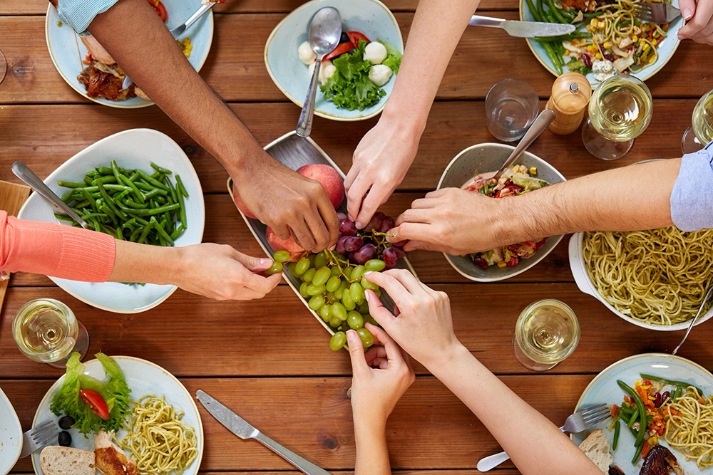Trebord med skåler av forskjellig type mat med en sirkel av armer i ulike hudfarger som griper tak i druer i midten av bordet.