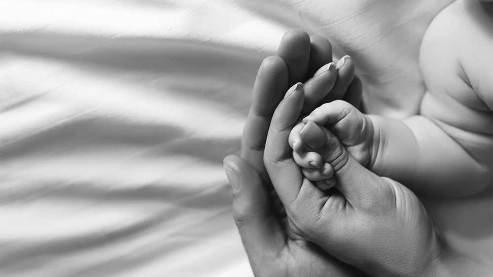 Et svart hvitt bilde av en to voksne hender som holder rundt hånden til et spedbarn.