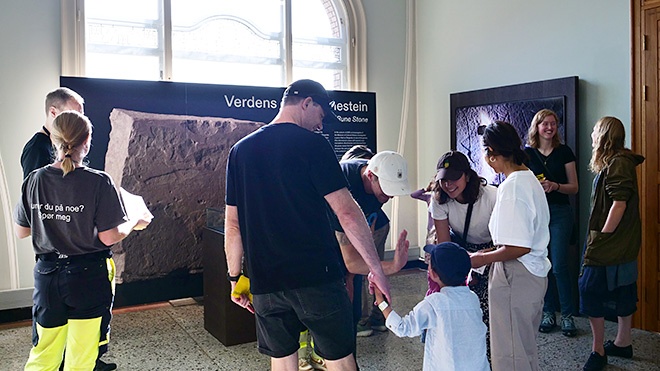 Barn og voksne står i et utstillingsrom og ser på et monter med en utstilt runestein.