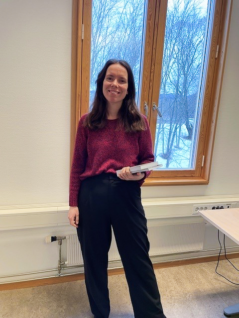 Astrid Helene Olsen står foran et vindu på kontoret sitt med en bunke papirer i ene armen. Hun smiler til kameraet.