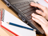 Hender som skriver på laptop, ved siden av notatblokk og blyant
