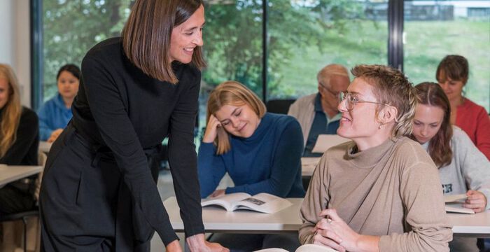 Undervisningsrom med studenter. Kvinnelig lærer står ved pulten til en elev og de smiler til hverandre. 