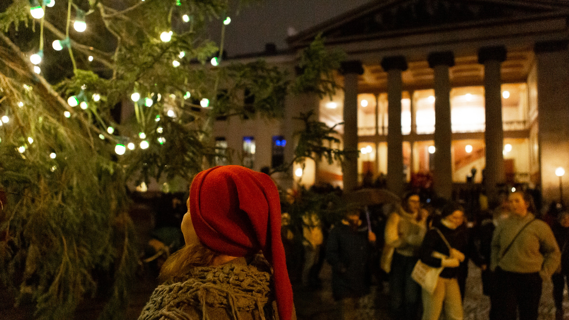 En kvinne står med ryggen til kameraet og har en rød nisselue på. Hun ser på juletreet på Universitetsplassen, som er tent med små lys. I bakgrunnen ser vi Universitetets aula.