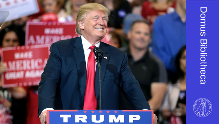 Donald Trump, en hvit mann i mørkeblå dressjakke og rødt slips, står på en talerstol og smiler. 