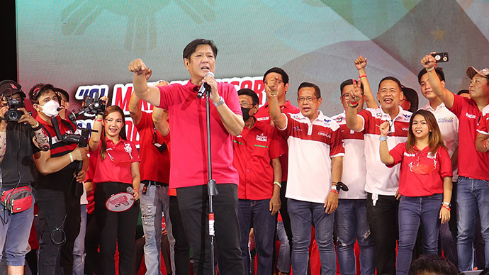 Asiatisk utseende mann i rød t-skjorte holder tale foran en gruppe mennesker. 