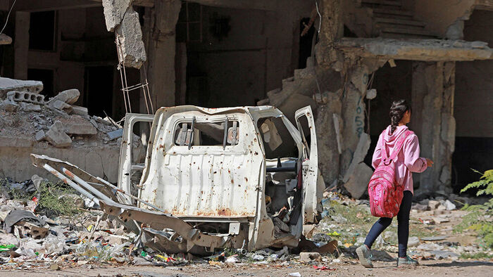En jente kledd i rosa går forbi et bilvrak blant ruiner.