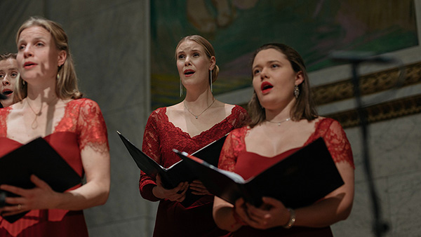 Tre sangere med notebøker i røde kjoler på scenen i Aulaen
