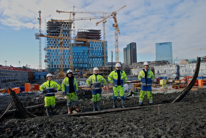 Fem arbeidere i selvlysende gule drakter med hjelm. I bakgrunnen ser vi flere høye blokker og fire-fem heisekraner.