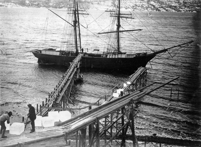 Avstandsbilde av en seilskute uten seil som ligger til kai. I forgrunnen to personer med fem-seks store isklumper. Sort-hvitt foto.