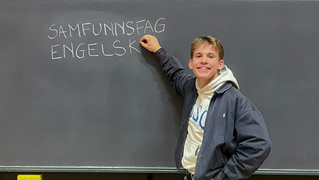 Student står foran tavle hvor han har skrevet "SAMFUNNSFAG" og "ENGELSK" med kritt.