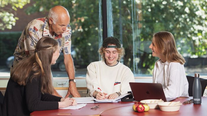 Tre studenter sitter ved et bord med PC og skrivesaker, og en foreleser står lent over bordet