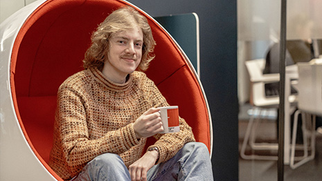 Mannlig student sitter i en stol og smiler skrått mot kamera mens han holder en kaffekopp i hånden