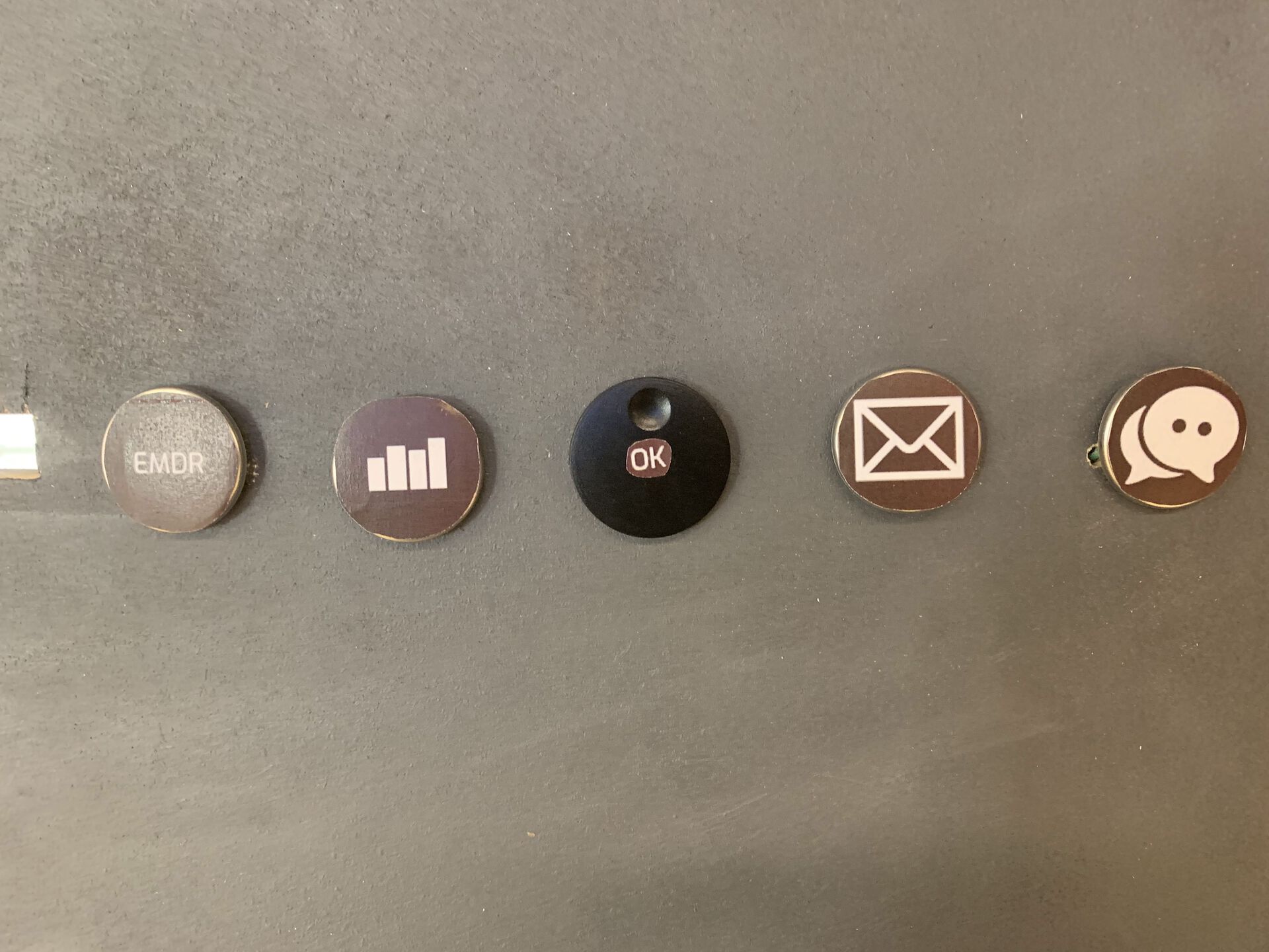 Et bilde av knappene på artefakten iRo. Fra venstre har man; EMDR-knapp, stemningsrapport-knapp, vrider med OK-knapp, sende rapport til ansatt-knapp, og samtale-knapp.