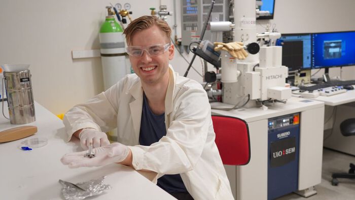 Student med labfrakk, vernebriller og med en nanoprøve i hånden. Sted: SEM-lab. 