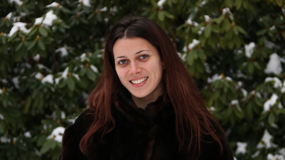 Ung kvinne med langt mørkt hår og svart jakke ser mot kamera og smiler, i bakgrunnen bartrær med snø. Foto.