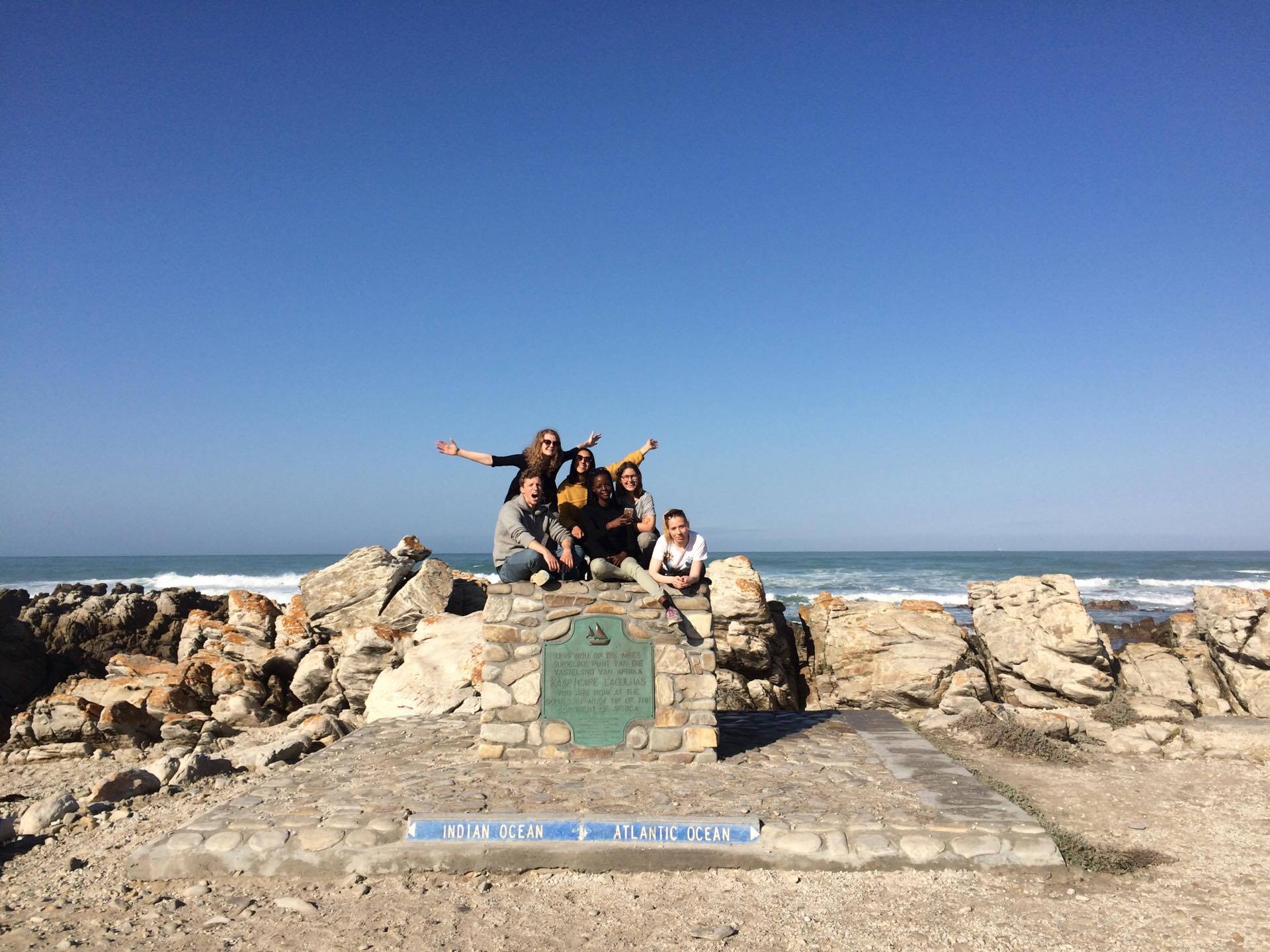 Flere personer som står på en sten som merker Afrikas Sørligste punkt, alle er glade og strekker armene ut i været