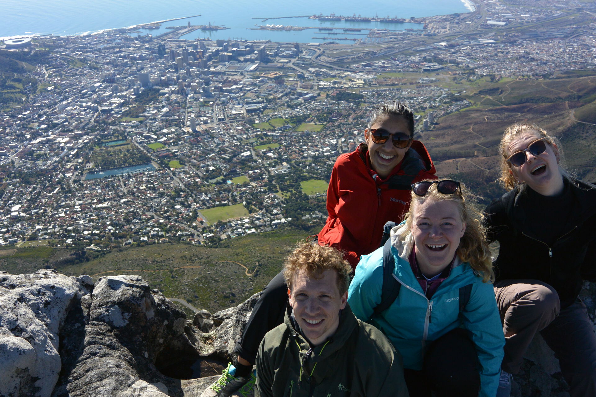 Fire personer som sitter på toppen av et fjell og smilier mot kamera med byen Cape Town i bakgrunnen