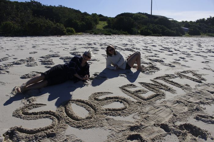 To jenter som ligger på stranden der det står skrevet i sanden SOSENT