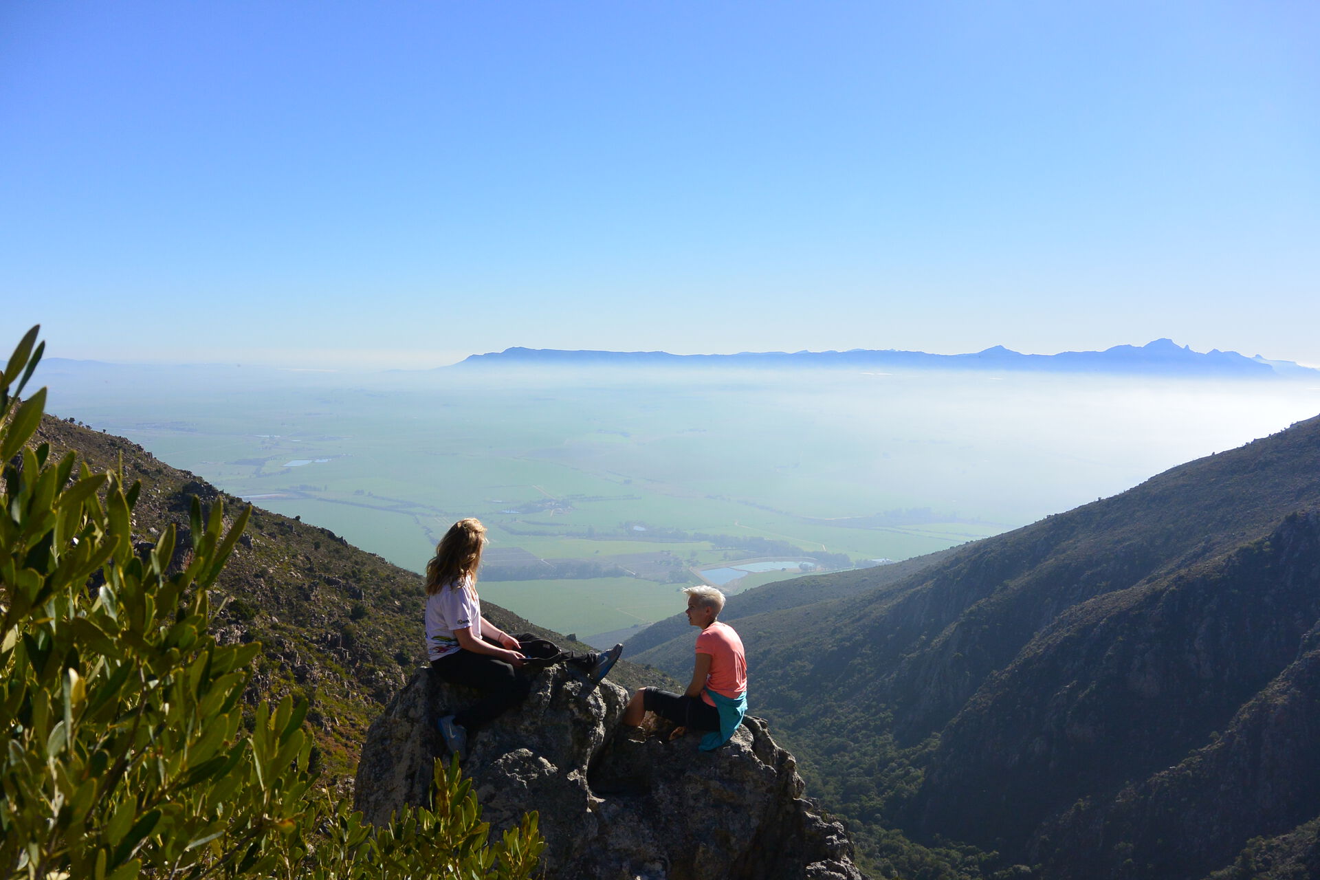 To jenter som sitter på toppen av et fjell med havet i bakgrunnen