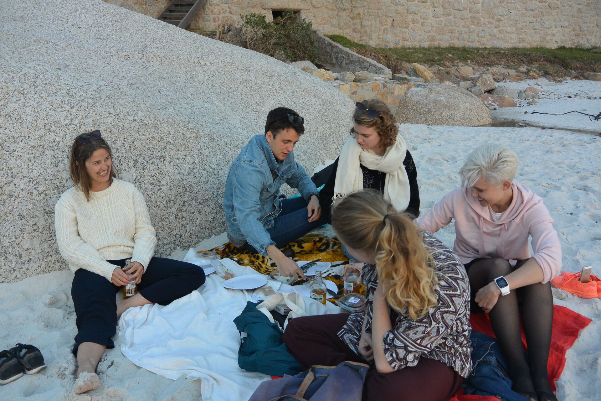 Fem unge voksne som sitter på stranden ved siden av en stein og har piknik.
