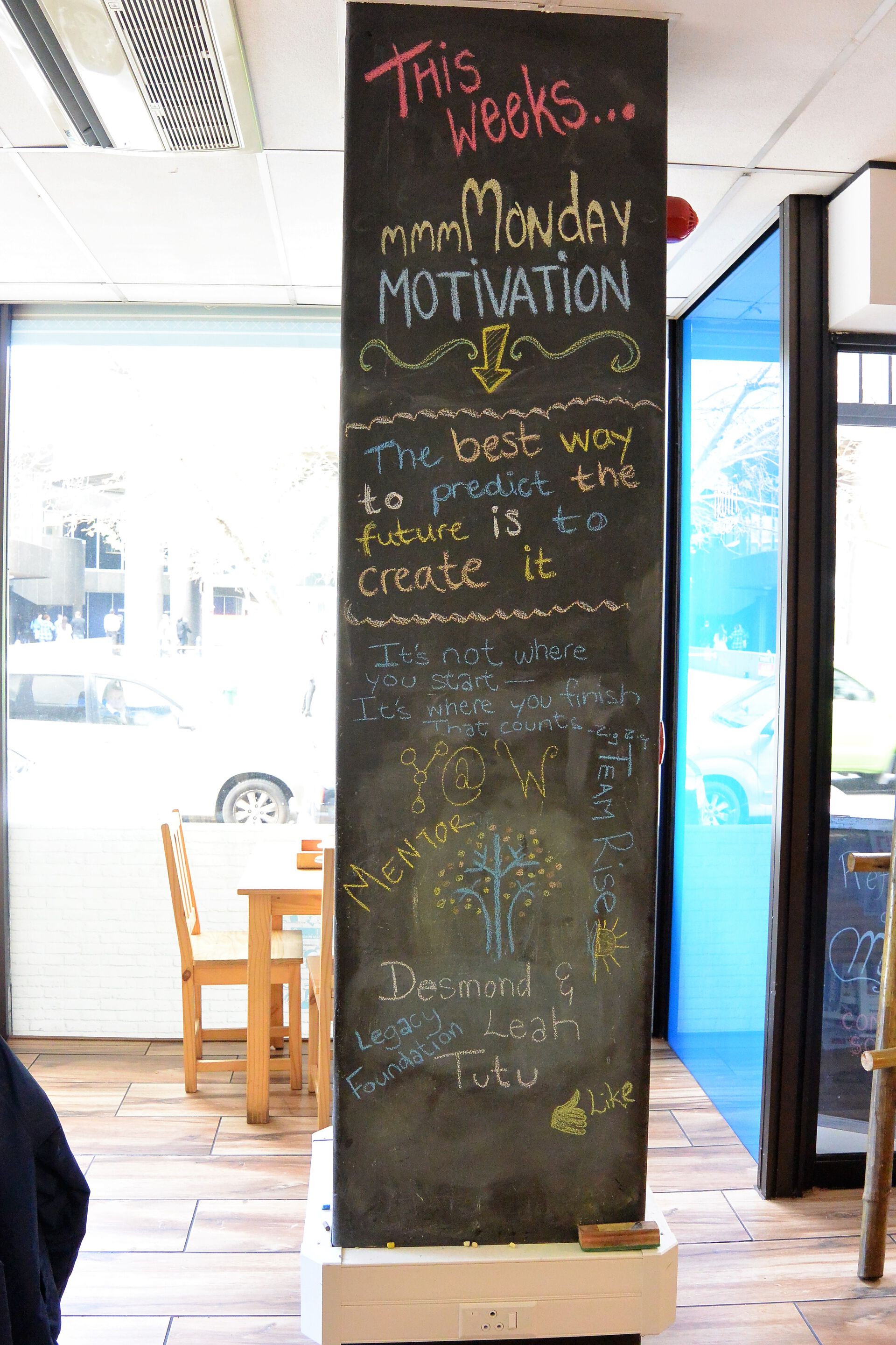En høy og smal tavle som står på gulvet i en kafe der det er skrevet med kritt i forskjellige farger motivasjonssitater