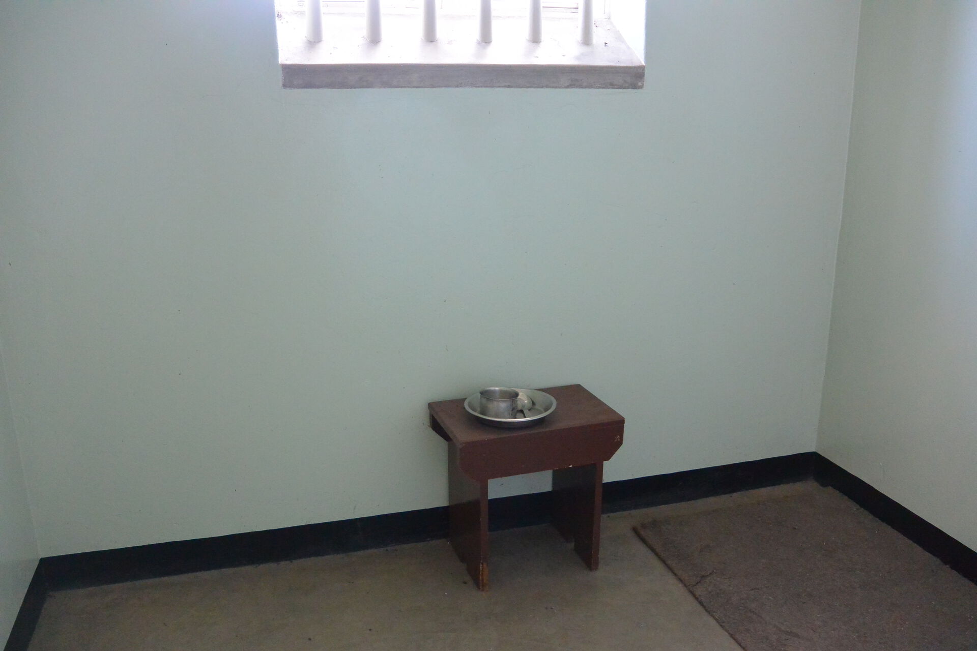 Et lite rom med vegger i mur og et vindu med gitter foran, en liten stol og et enkelt teppe.